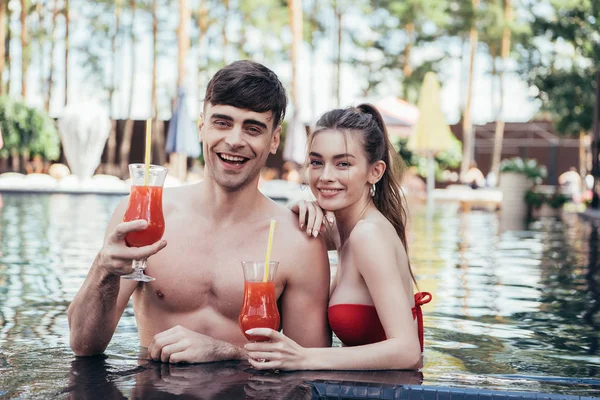 Feliz joven pareja sonriendo a la cámara mientras se relaja en la piscina con vasos de bebida refrescante - foto de stock