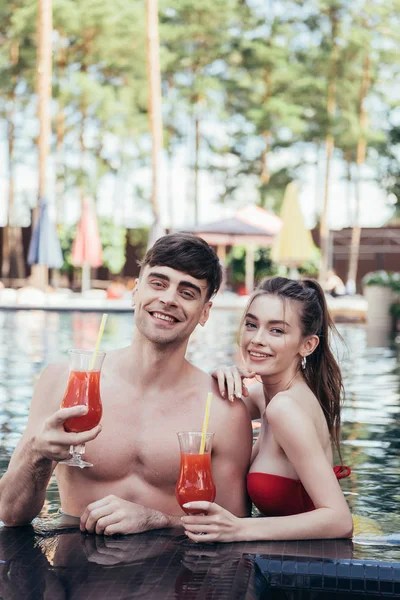 Alegre joven pareja sonriendo a la cámara mientras se relaja en la piscina con vasos de bebida refrescante - foto de stock