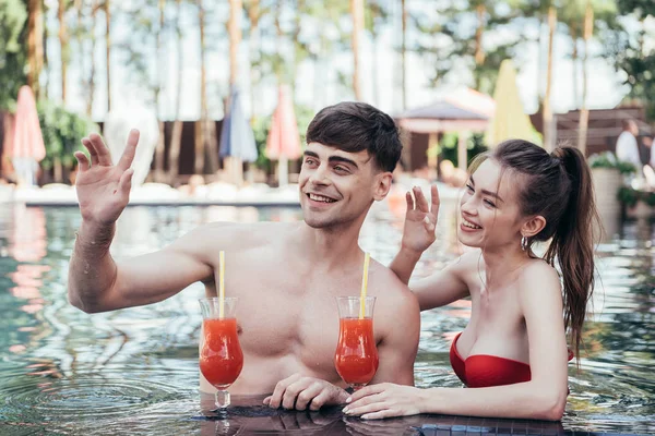 Alegre pareja joven mirando hacia otro lado y agitando las manos mientras se relaja en la piscina con vasos de bebida refrescante - foto de stock