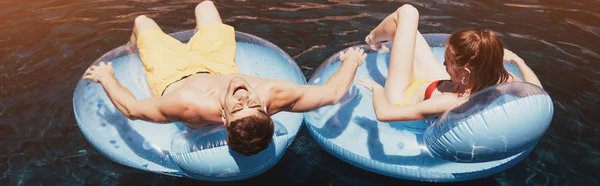 Tiro panorâmico de banho de sol jovem casal em anéis de natação na piscina — Fotografia de Stock
