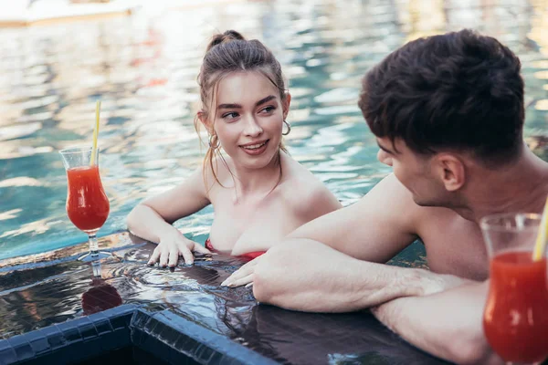 Felice giovane coppia che si guarda mentre riposa a bordo piscina con bicchieri di bevanda rinfrescante — Foto stock