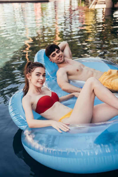 Счастливая молодая пара, плавающая на плавательных кольцах в бассейне и улыбающаяся перед камерой — стоковое фото