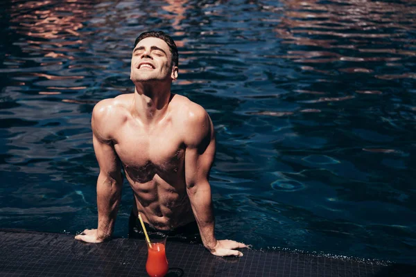 Bel homme joyeux relaxant au bord de la piscine près d'un verre de boisson rafraîchissante — Photo de stock