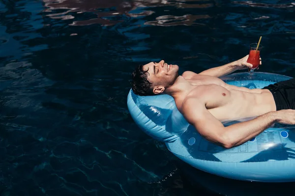 Щасливий молодий чоловік розслабляється на плавальному кільці в басейні зі склянкою освіжаючого напою — стокове фото