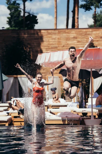 Excitada pareja joven cogida de la mano mientras salta en la piscina - foto de stock