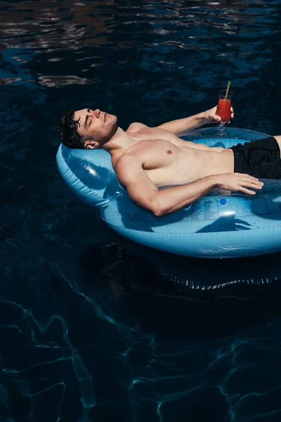 Joven guapo sosteniendo un vaso de bebida refrescante mientras flota en el anillo de natación en la piscina - foto de stock