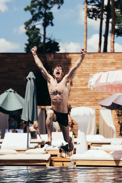 Excited young man screaming while jumping in swimming pool with raised hands — Stock Photo