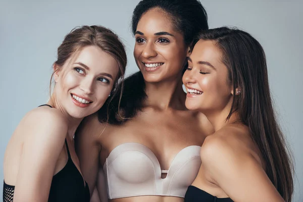 Три сексуальные многоэтнические девушки в нижнем белье смеются и обнимаются изолированно на серой — стоковое фото