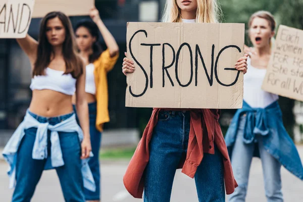 Vista cortada de cartaz feminista segurando com inscrição forte perto de mulheres na rua — Fotografia de Stock