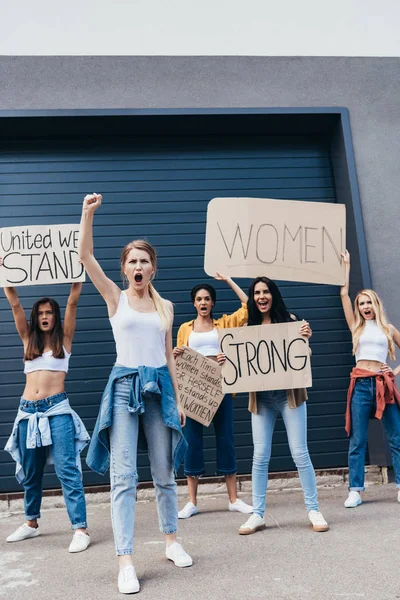 Vista completa de feministas multiétnicas gritando sosteniendo pancartas con consignas en la calle - foto de stock