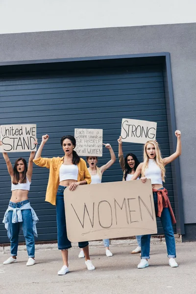 Vista completa de feministas multiétnicas gritando sosteniendo pancartas con consignas en la calle - foto de stock