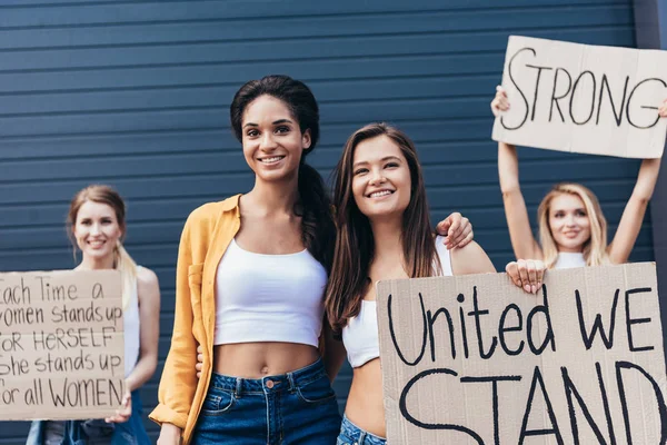 Vue de face des féministes souriantes multiethniques embrassant et tenant la pancarte avec l'inscription unie nous nous tenons dans la rue — Photo de stock