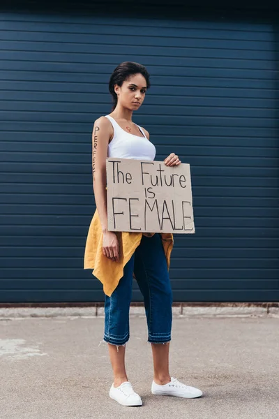 Vista completa de feminista afroamericana con inscripción perfecta de brazo sosteniendo pancarta con lema el futuro es femenino en la calle - foto de stock