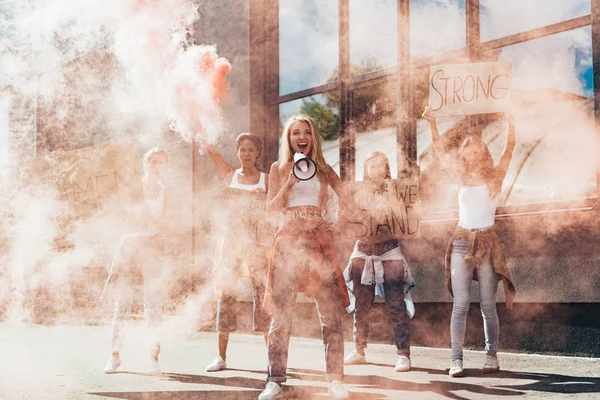 Vista completa de feministas multiétnicas gritando con carteles de altavoces con eslóganes en humo rojo en la calle - foto de stock