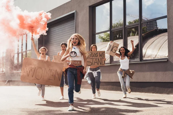 Vista completa de feministas multiétnicas gritando con carteles de altavoces con consignas y corriendo por la calle - foto de stock