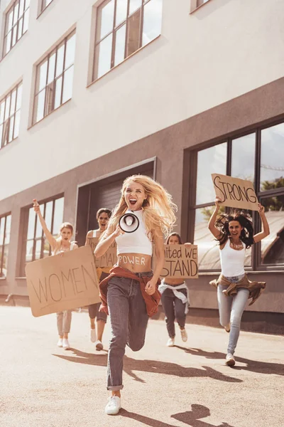Vista completa de feministas gritando con altavoces sosteniendo pancartas con consignas y corriendo por la calle - foto de stock