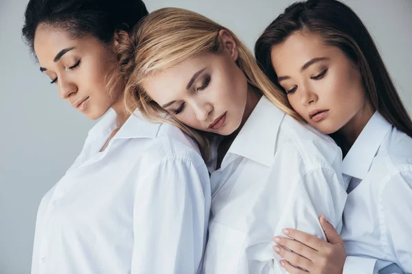 Tres atractivas chicas multiétnicas en camisas blancas abrazando aisladas en gris - foto de stock