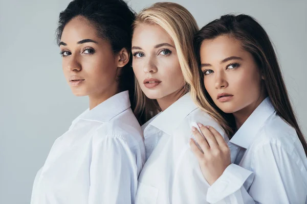 Tres atractivas chicas multiétnicas en camisas blancas abrazando aisladas en gris - foto de stock