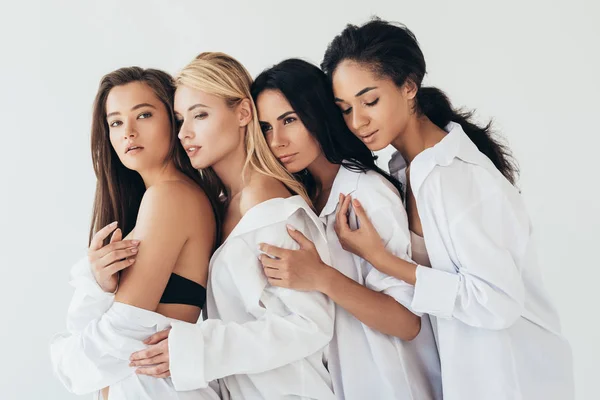 Cuatro atractivas mujeres multiétnicas en camisas blancas abrazando aisladas en gris - foto de stock