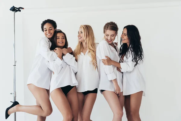 Sexy multiethnic feminists wearing white shirts smiling and embracing on grey — Stock Photo