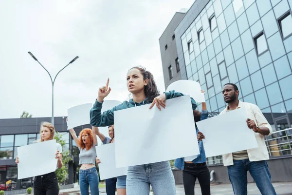 Fuoco selettivo della donna attraente che indica con il dito mentre tiene il cartello vuoto vicino alla gente multiculturale — Foto stock