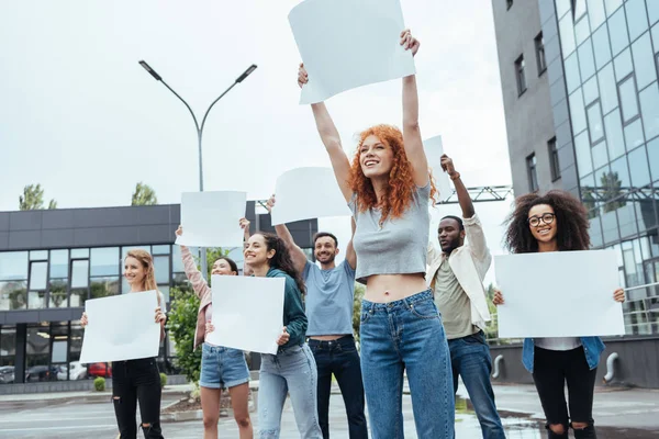 Selective focus of redhead girl holding empty board near multicultural people — Stock Photo