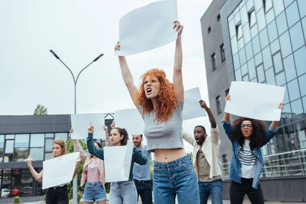 Selective focus of angry redhead girl holding blank placard on meeting near multicultural people — Stock Photo