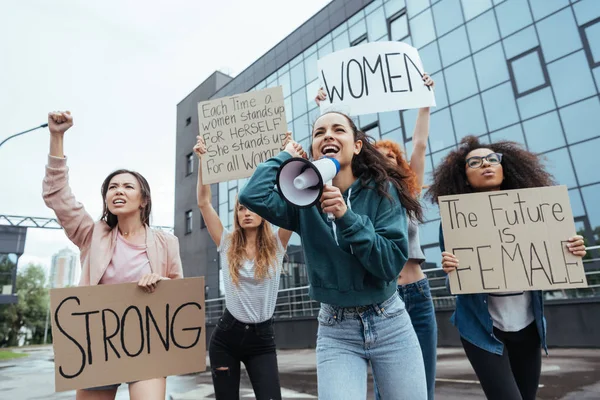 Foco seletivo de menina emocional segurando megafone e gritando perto de mulheres multiculturais segurando cartazes em reunião — Fotografia de Stock