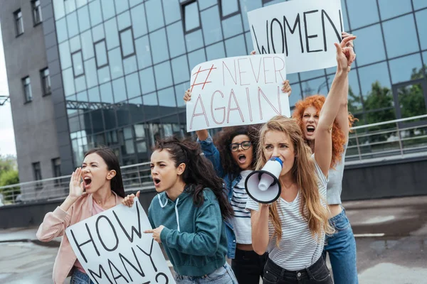 Selective focus of emotional girl holding megaphone and gesturing near multicultural women holding placards on meeting — Stock Photo