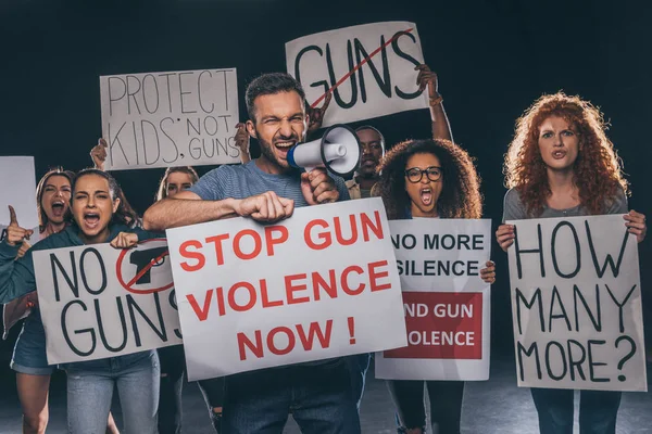Uomo arrabbiato tenendo cartello con fermare la violenza pistola ora lettering e urlando in megafono vicino a persone multiculturali su nero — Foto stock