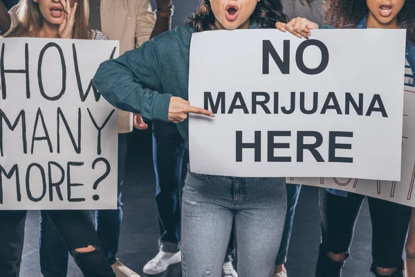 Обрезанный вид эмоциональной девушки, указывающей пальцем на плакат без марихуаны, здесь, рядом с мультикультурными людьми на черном — стоковое фото