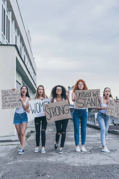 Эмоциональные мультикультурные девушки, держащие плакаты и кричащие возле здания — стоковое фото