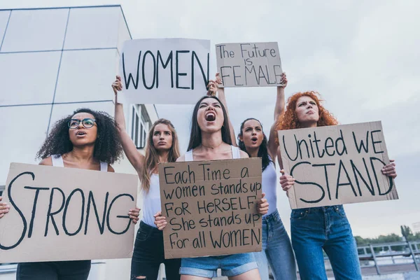 Vue en angle bas de femmes multiculturelles tenant des pancartes et criant à l'extérieur — Photo de stock