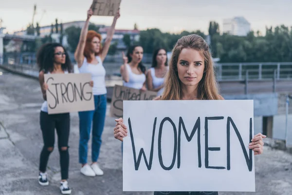 Selective focus of woman holding board with women lettering near multicultural girls outside — Stock Photo