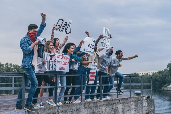 Hombre con bufanda en la cara gritando en megáfono cerca de personas multiculturales con letras en carteles cerca del río - foto de stock