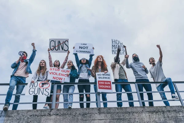 Vista de ángulo bajo del hombre con bufanda en la cara gritando en megáfono cerca de personas multiculturales con letras en pancartas haciendo gestos fuera - foto de stock