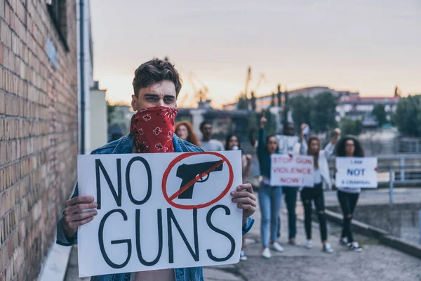 Foco seletivo do homem com cachecol no rosto segurando cartaz sem armas lettering perto de pessoas multiculturais — Fotografia de Stock