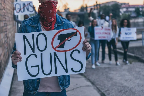 Vista recortada del hombre con bufanda en la cara con pancarta sin letras de armas cerca de personas multiculturales - foto de stock