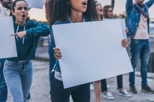 Vista recortada de chicas emocionales multiculturales gritando y sosteniendo pancartas en blanco durante la protesta - foto de stock
