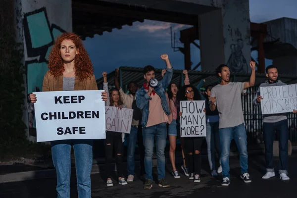 Enfoque selectivo de chica pelirroja sosteniendo cartel con mantener a los niños letras seguras cerca de personas multiculturales en el encuentro - foto de stock