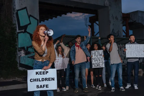 Селективное внимание эмоциональной рыжей девушки, держащей плакат с безопасными надписями и кричащей рядом с мультикультурными людьми на встрече — стоковое фото