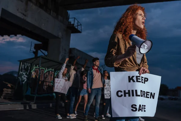 Селективный фокус эмоциональной рыжей девушки с плакатом с сохранением безопасности детей рядом с мультикультурными людьми на встрече — стоковое фото