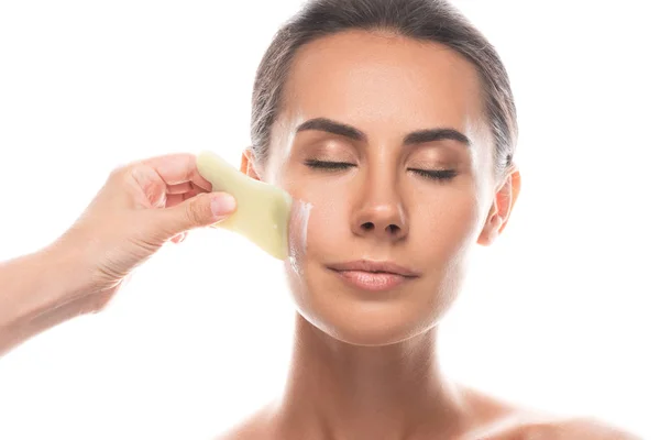 Vue partielle du cosmétologue appliquant la crème pour le visage et utilisant gua sha grattoir isolé sur blanc — Photo de stock