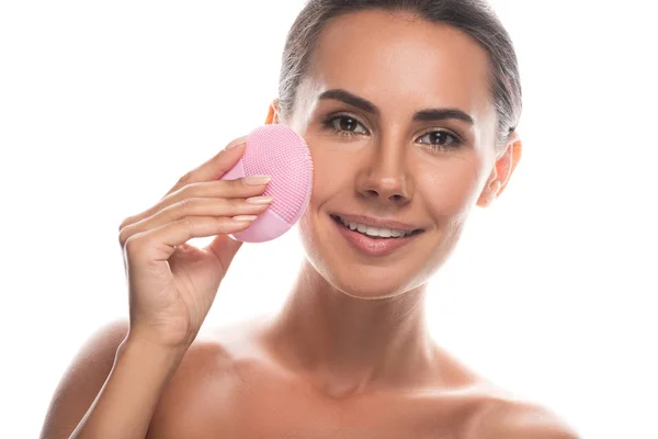 Vue de face de sourire jeune femme nue à l'aide d'une brosse nettoyante faciale isolée sur blanc — Photo de stock