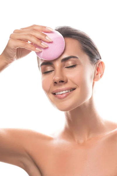 Sonriente mujer joven desnuda usando cepillo de limpieza facial con los ojos cerrados aislados en blanco - foto de stock