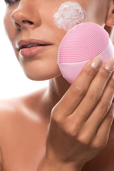 Vista cortada de mulher jovem nua com creme no rosto usando escova de limpeza facial isolada em branco — Fotografia de Stock