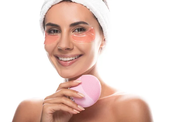 Smiling nude young woman with eye patches holding facial cleansing brush isolated on white — Stock Photo