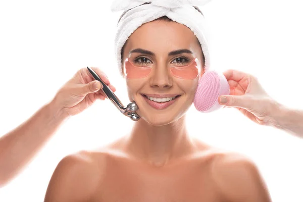 Cropped view of cosmetologists using facial cleansing brush and facial massager and smiling woman with eye patches isolated on white — Stock Photo