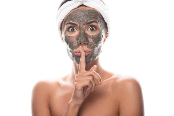 Vue de face de surprise jeune femme nue en bande capillaire cosmétique avec masque en argile sur le visage montrant signe de silence isolé sur blanc — Photo de stock