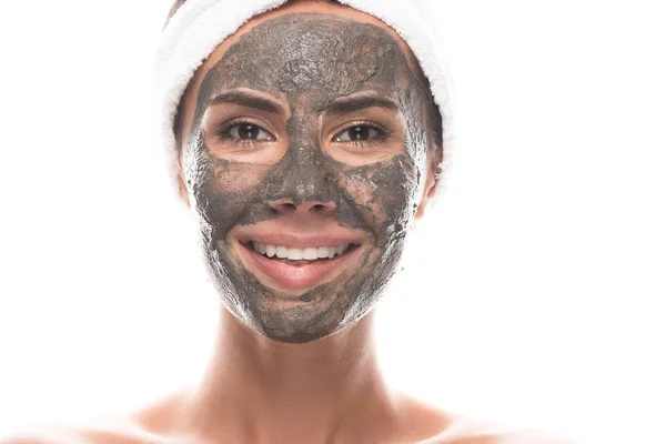 Vue de face de jeune femme nue souriante en bande capillaire cosmétique avec masque en argile sur le visage isolé sur blanc — Photo de stock